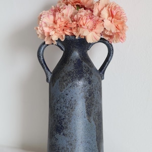 Op de afbeelding: Een donkerblauwe keramische vaas met twee handvatten, gevuld met perzikroze anjers. De vaas heeft een getextureerd, gevlekt uiterlijk. De bloemen zijn aan de bovenkant gegroepeerd, wat een contrast vormt met de vaas.