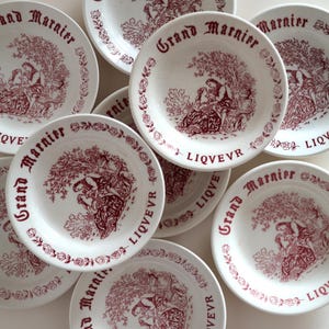 Peut inclure: Plusieurs assiettes en céramique blanche avec une illustration et un texte bordeaux. Chaque assiette présente une scène détaillée de personnages sous un arbre et les mots "Grand Marnier" et "LIQVEVR". Les assiettes sont décorées d'une bordure florale.