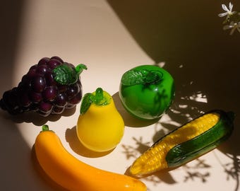 May include: A collection of five glass fruit and vegetable figurines including a cluster of dark purple grapes, a bright yellow pear, a vibrant green apple, a smooth yellow banana, and a yellow corn cob encased in dark green glass husks.