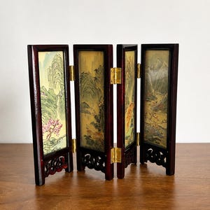 May include: A four-panel folding screen featuring a dark wood frame with intricate carved bases and gold-colored metal hinges. Each panel displays a painted landscape scene with mountains, trees, and pink blossoms on a light-colored background.