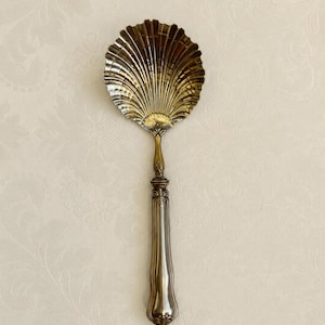 Peut inclure: Une cuillère de service argentée dotée d'un cuilleron en forme de coquille Saint-Jacques avec des rainures profondes. Le manche est allongé et lisse, avec une jonction métallique ornementée et travaillée, se terminant par une base arrondie.