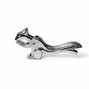May include: A silver-colored metal nutcracker shaped like a squirrel. The tool features a hinged jaw with serrated teeth for gripping and a long, curved tail that serves as the handle for applying pressure to crack open nuts.