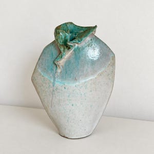 Può includere: Un vaso in ceramica dalla silhouette asimmetrica e angolare. La parte inferiore presenta uno smalto bianco sporco maculato, mentre la sezione superiore e il bordo ripiegato e ondulato sono rifiniti con uno smalto blu turchese lucido.