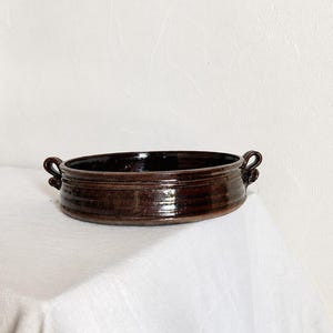 Peut inclure: Un plat de cuisson rond en céramique émaillée brun foncé avec une finition brillante. Le plat présente un bassin large et peu profond ainsi que deux petites anses décoratives en boucle sur les côtés, avec des stries horizontales.