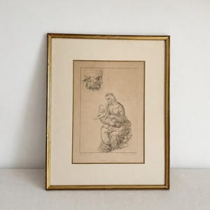 Peut inclure: Une impression encadrée représentant un croquis d'une personne tenant un enfant, avec un croquis séparé d'une tête au-dessus. L'œuvre est réalisée en traits noirs sur fond beige, insérée dans un cadre doré fin avec un large passe-partout crème.
