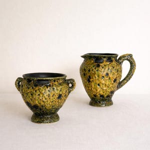 May include: Two ceramic vessels with a textured, speckled design in shades of green, yellow, and black. One is a small vase with handles, and the other is a pitcher with a handle and spout. Both have a dark rim.