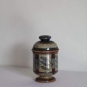 May include: A ceramic jar with a lid, featuring a speckled beige and blue glaze. The jar has a cylindrical body with a pedestal base and is adorned with a repeating pattern of blue and brown leaf-like shapes. The lid is dark blue.