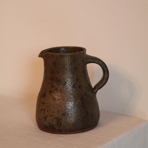 Puede incluir: Jarra de cerámica marrón oscuro hecha a mano, con superficie texturizada y un pequeño pico. La jarra tiene un cuerpo redondeado, un asa robusta y una base de color marrón rojizo. Esta pieza rústica es adecuada para servir bebidas.