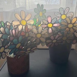 May include: Two decorative flower arrangements in pots. The flowers are made of stained glass in various colours, including yellow, pink, blue, and green. The pots are different colours and patterns. The flowers are arranged to resemble a bouquet.