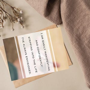 May include: A greeting card with the text "I AM EASILY MANIFESTING AND CONFIDENTLY STEPPING INTO THE LIFE I'VE ALWAYS ENVISIONED." The card is on a brown envelope, with a beige and brown background. A sprig of dried flowers and a brown cloth are also in the image.