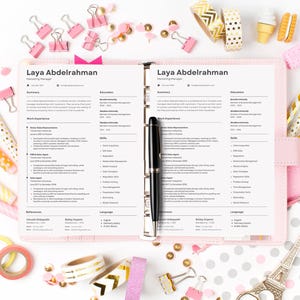 May include: A pink planner displaying a resume titled "Laya Abdelrahman" is surrounded by stationery. The desk is decorated with pink paper clips, washi tape, pencils, and decorative erasers. A black pen rests on the resume.