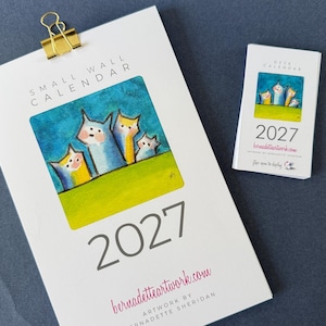 May include: A 2027 small wall calendar and a matching desk calendar. Both feature a colorful illustration of four stylized figures against a blue sky and green ground. The wall calendar has a gold binder clip at the top and text reading 2027.