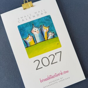 May include: A 2027 small wall calendar featuring a gold metal binder clip at the top. The cover displays a colorful painting of four stylized cat-like figures against a blue sky and green grass, with the text bernadetteartwork.com and Bernadette Sheridan.