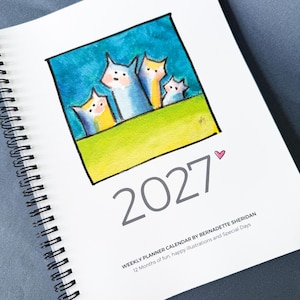 May include: A spiral-bound 2027 weekly planner calendar featuring a watercolor illustration of four stylized cats against a blue sky and green field. The cover includes a small pink heart, the text 2027, and the title Weekly Planner Calendar by Bernadette Sheridan, 12 Months of fun, happy illustrations and Special Days.