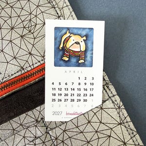 May include: A silver wallet with a geometric black line pattern and an orange zipper, holding a white calendar card for April 2027. The card features a watercolor illustration of a tan bulldog wearing a striped sweater against a blue background.