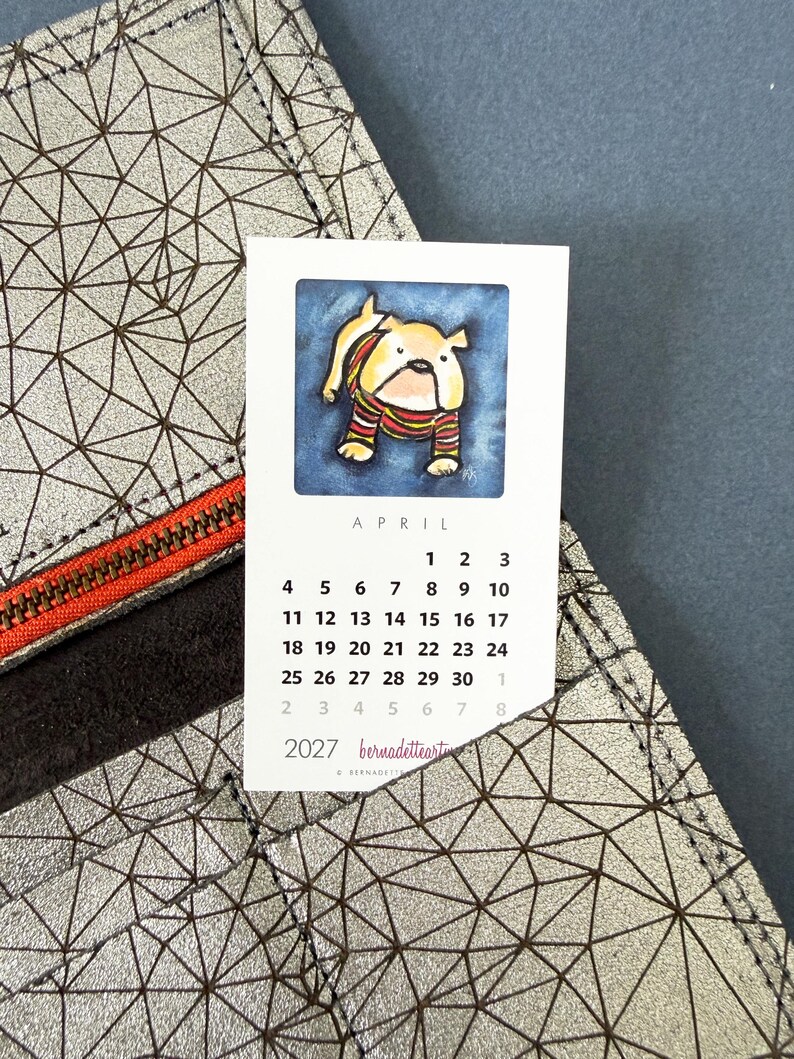 May include: A silver wallet with a geometric black line pattern and an orange zipper, holding a white calendar card for April 2027. The card features a watercolor illustration of a tan bulldog wearing a striped sweater against a blue background.