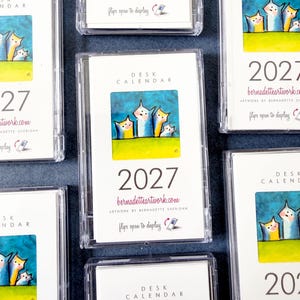 May include: Multiple 2027 desk calendars in clear plastic cases. Each white card features a colorful illustration of four stylized cats against a blue sky and green grass, with the text 'DESK CALENDAR', '2027', and 'bernadetteartwork.com'.