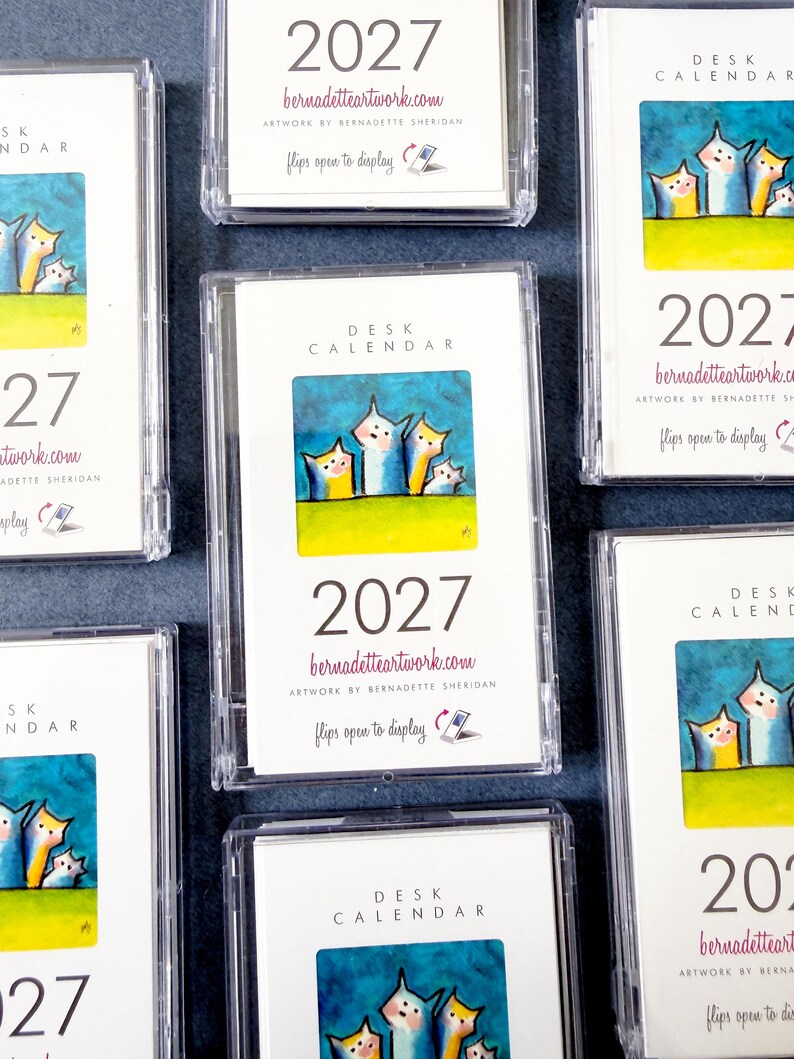 May include: Multiple 2027 desk calendars in clear plastic cases. Each white card features a colorful illustration of four stylized cats against a blue sky and green grass, with the text 'DESK CALENDAR', '2027', and 'bernadetteartwork.com'.