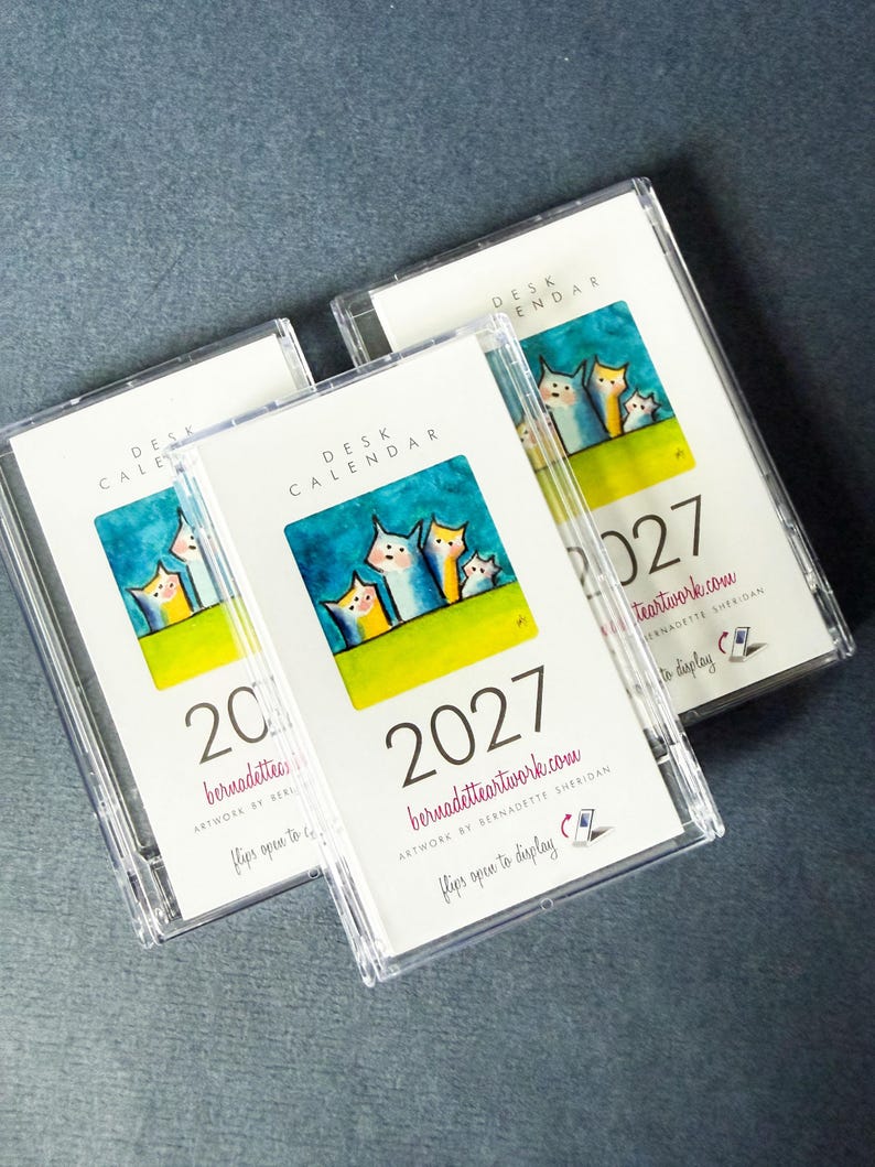May include: Three clear plastic cases containing 2027 desk calendars. Each calendar features a white cover with the text DESK CALENDAR, the year 2027, the website bernadetteartwork.com, and a colorful illustration of three stylized cats.