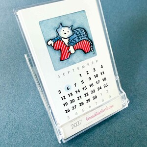 May include: A 2027 desk calendar in a clear acrylic stand featuring a watercolor illustration of a cat in a blue sweater resting on a red and white striped chair. The page shows the month of September with a blue circle around the number 6.