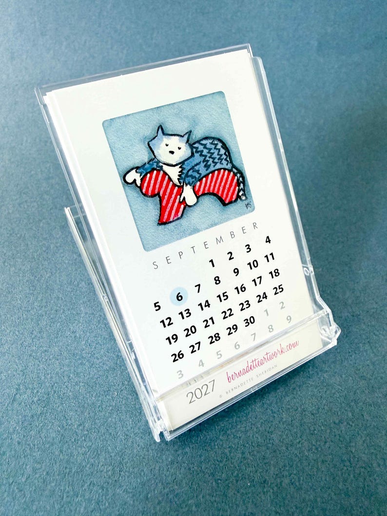 May include: A 2027 desk calendar in a clear acrylic stand featuring a watercolor illustration of a cat in a blue sweater resting on a red and white striped chair. The page shows the month of September with a blue circle around the number 6.