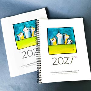 May include: Two white spiral-bound weekly planner calendars for 2027. The cover features a square illustration of four stylized cats against a blue sky and green field. Text reads: 2027, WEEKLY PLANNER CALENDAR BY BERNADETTE SHERIDAN, 12 Months of fun.