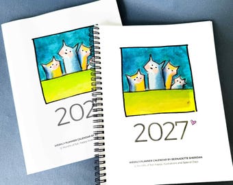 May include: Two white spiral-bound weekly planner calendars for 2027. The cover features a square illustration of four stylized cats against a blue sky and green field. Text reads: 2027, WEEKLY PLANNER CALENDAR BY BERNADETTE SHERIDAN, 12 Months of fun.