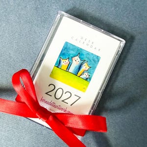 May include: A 2027 desk calendar encased in a clear plastic cassette-style case, tied with a bright red satin ribbon. The cover features a watercolor illustration of four stylized cats against a blue sky and green grass, with the text 2027.