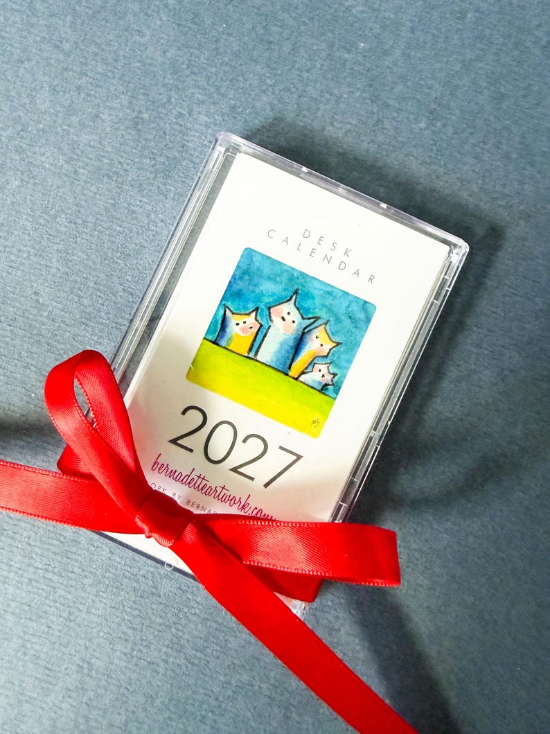 May include: A 2027 desk calendar encased in a clear plastic cassette-style case, tied with a bright red satin ribbon. The cover features a watercolor illustration of four stylized cats against a blue sky and green grass, with the text 2027.