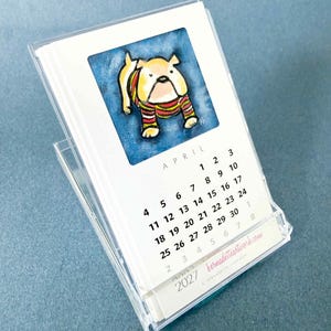 May include: A 2027 desk calendar in a clear acrylic stand featuring a watercolor illustration of a bulldog wearing a striped sweater. The page shows the month of April with black text on a white background and the website bernadetteartwork.com.