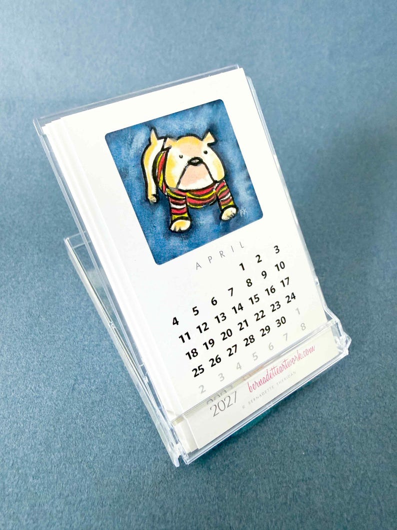 May include: A 2027 desk calendar in a clear acrylic stand featuring a watercolor illustration of a bulldog wearing a striped sweater. The page shows the month of April with black text on a white background and the website bernadetteartwork.com.