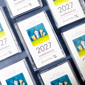 May include: Multiple 2027 desk calendars in clear plastic cases. Each features a colorful illustration of four stylized cats against a blue sky and green grass. Text includes 'DESK CALENDAR', '2027', 'bernadetteartwork.com', and 'flips open to display'.