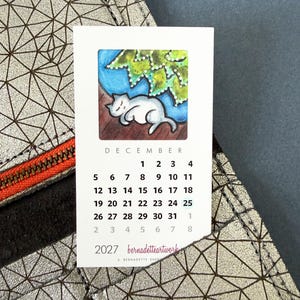 May include: A rectangular 2027 calendar card for December featuring a white cat sleeping under green leaves. The card displays the month, dates, and the text 'bernadetteartwork' and 'BERNADETTE SHERRY'. It is placed against a metallic silver geometric patterned surface.