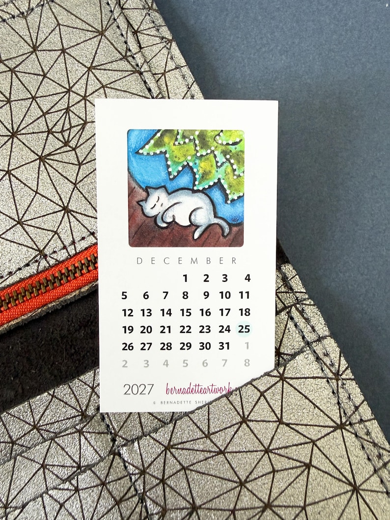 May include: A rectangular 2027 calendar card for December featuring a white cat sleeping under green leaves. The card displays the month, dates, and the text 'bernadetteartwork' and 'BERNADETTE SHERRY'. It is placed against a metallic silver geometric patterned surface.