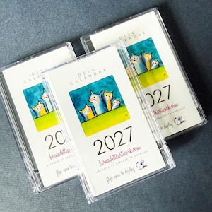 May include: Three clear plastic cases containing 2027 desk calendars. Each calendar features a white cover with the text DESK CALENDAR, the year 2027, the website bernadetteartwork.com, and a colorful illustration of three stylized cats.