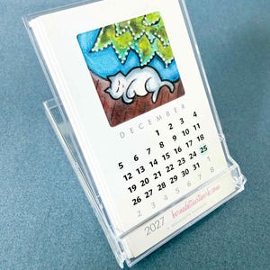 May include: A 2027 desk calendar in a clear acrylic stand featuring a watercolor illustration of a white cat sleeping under a green tree. The page shows the month of December with black text and the website bernadetteartwork.com printed at the bottom.