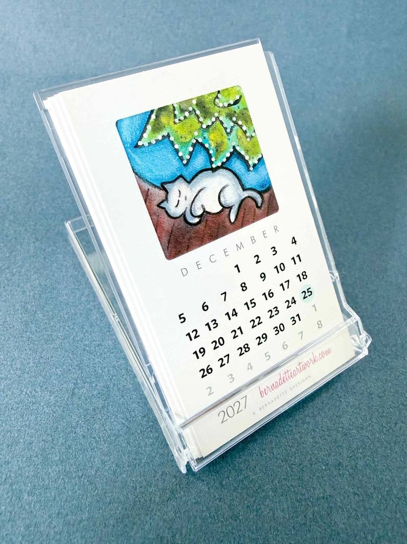 May include: A 2027 desk calendar in a clear acrylic stand featuring a watercolor illustration of a white cat sleeping under a green tree. The page shows the month of December with black text and the website bernadetteartwork.com printed at the bottom.