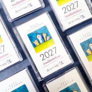 May include: A collection of 2027 desk calendars in clear plastic cases. Each calendar features a colorful illustration of four stylized cats on a green field against a blue background, with the text 'DESK CALENDAR', '2027', and 'bernadetteartwork.com'.