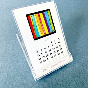 May include: A 2027 desk calendar in a clear acrylic stand featuring a July page with a square graphic of vertical stripes in red, orange, yellow, green, blue, and purple. The calendar displays the month, dates, and the website bernadetteartwork.com.