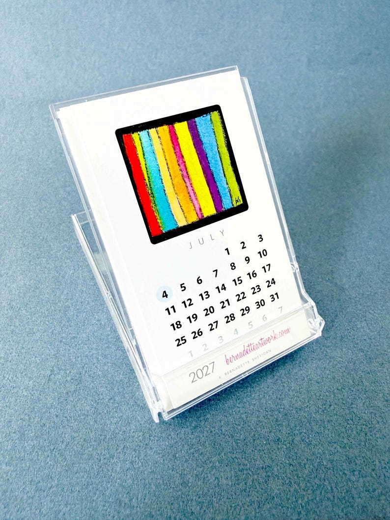 May include: A 2027 desk calendar in a clear acrylic stand featuring a July page with a square graphic of vertical stripes in red, orange, yellow, green, blue, and purple. The calendar displays the month, dates, and the website bernadetteartwork.com.