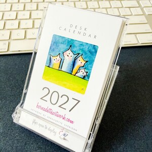 May include: A 2027 desk calendar in a clear plastic case. The cover features the text DESK CALENDAR, 2027, bernadetteartwork.com, and flips open to display. A square watercolor illustration shows four stylized cat-like figures on a green hill.