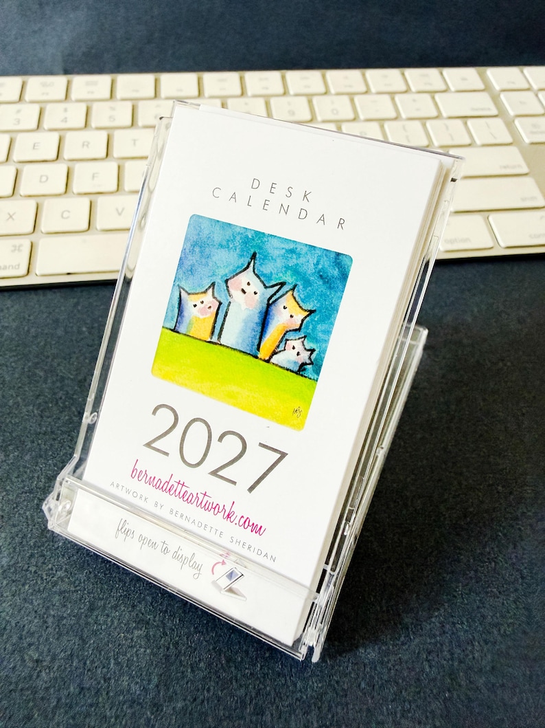 May include: A 2027 desk calendar in a clear plastic case. The cover features the text DESK CALENDAR, 2027, bernadetteartwork.com, and flips open to display. A square watercolor illustration shows four stylized cat-like figures on a green hill.