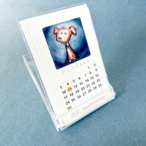 May include: A 2027 desk calendar in a clear plastic case featuring a watercolor illustration of a brown dog on a blue background for the month of October. The calendar displays the month name, dates, and the website bernadetteartwork.com.
