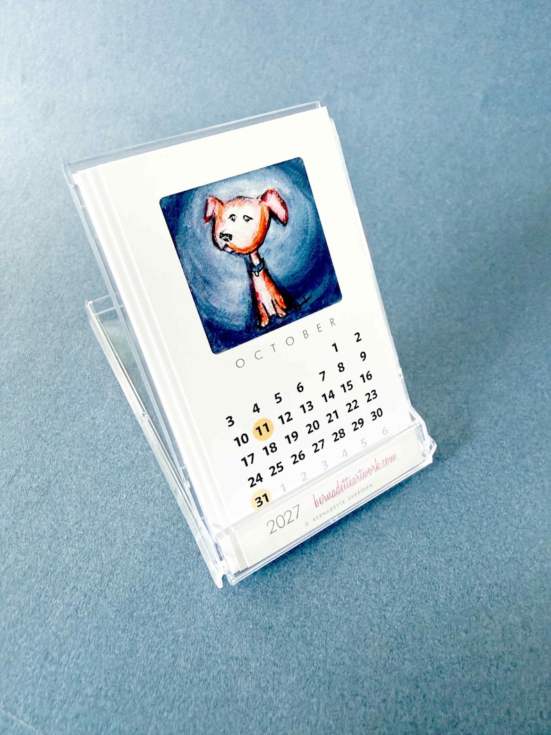 May include: A 2027 desk calendar in a clear plastic case featuring a watercolor illustration of a brown dog on a blue background for the month of October. The calendar displays the month name, dates, and the website bernadetteartwork.com.