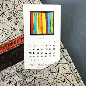 May include: A 2027 calendar card for July featuring a colorful vertical striped square graphic. The card is tucked into a silver metallic wallet with a dark brown geometric triangular pattern and a bright orange zipper detail.