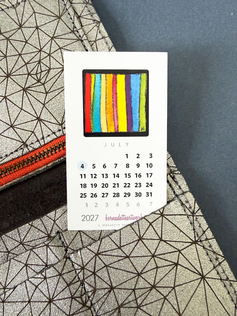 May include: A 2027 calendar card for July featuring a colorful vertical striped square graphic. The card is tucked into a silver metallic wallet with a dark brown geometric triangular pattern and a bright orange zipper detail.