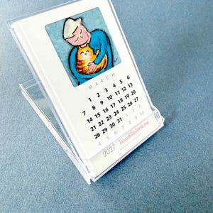 May include: A 2027 desk calendar in a clear acrylic stand featuring a March page with a watercolor illustration of a person in a blue shirt holding an orange cat. The calendar displays the month, dates, and the website bernadetteartwork.com.