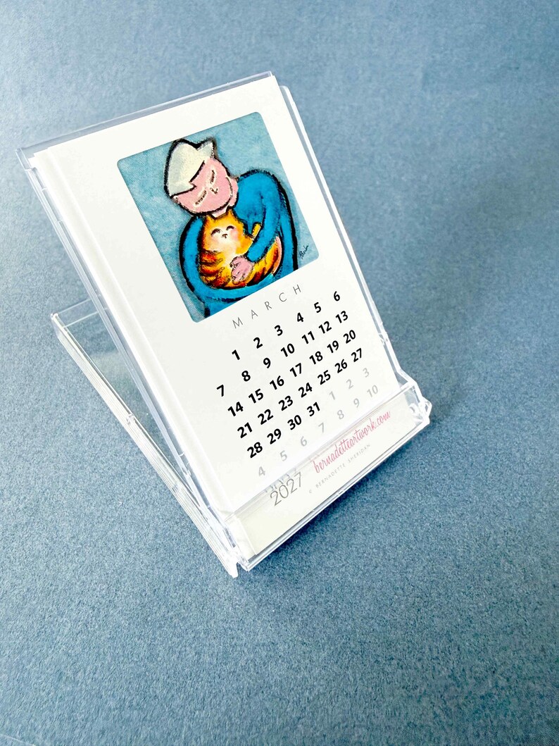May include: A 2027 desk calendar in a clear acrylic stand featuring a March page with a watercolor illustration of a person in a blue shirt holding an orange cat. The calendar displays the month, dates, and the website bernadetteartwork.com.