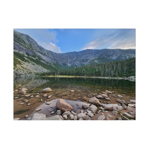May include: A scenic photograph of a mountain range reflected in a tranquil lake. The foreground shows clear water with visible rocks. The sky is blue with scattered clouds, and the mountains are covered in green vegetation.