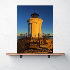 May include: A white lighthouse with a black top stands tall against a clear blue sky. The lighthouse is on a stone foundation with a metal fence and wooden stairs leading up to it. The image is a landscape photo.